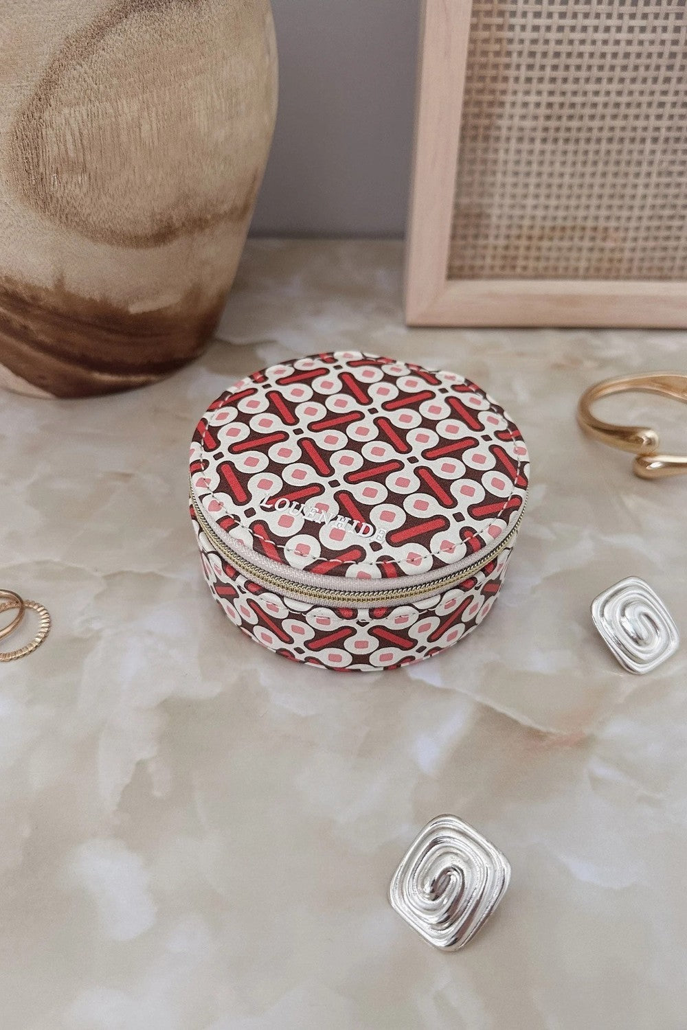 A small round zippered jewelry box with a red, white, and black geometric pattern, styled on a marble surface with gold and silver spiral earrings and a ceramic vase in the background.