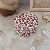 A small round zippered jewelry box with a red, white, and black geometric pattern, styled on a marble surface with gold and silver spiral earrings and a ceramic vase in the background.
