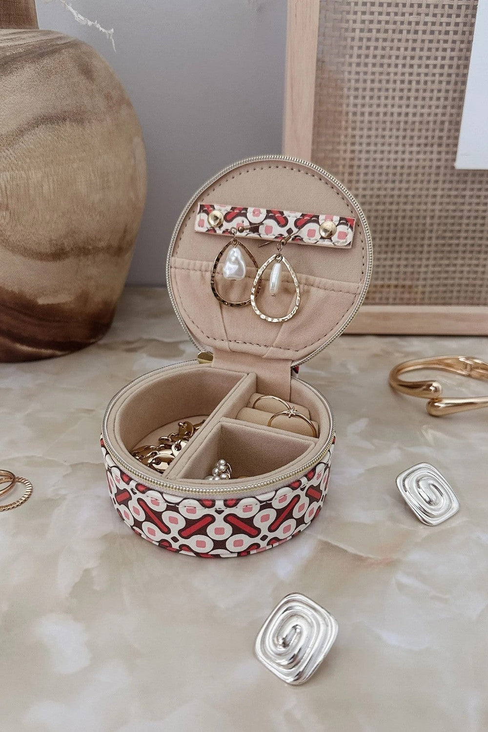 A round jewelry organizer case with a red, white, and pink pattern exterior and beige interior, shown open with earrings and rings inside. The case is styled on a beige marble-like surface with decorative jewelry pieces and a neutral background.