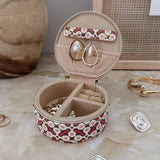 A round jewelry organizer case with a red, white, and pink pattern exterior and beige interior, shown open with earrings and rings inside. The case is styled on a beige marble-like surface with decorative jewelry pieces and a neutral background.
