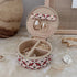 A round jewelry organizer case with a red, white, and pink pattern exterior and beige interior, shown open with earrings and rings inside. The case is styled on a beige marble-like surface with decorative jewelry pieces and a neutral background.