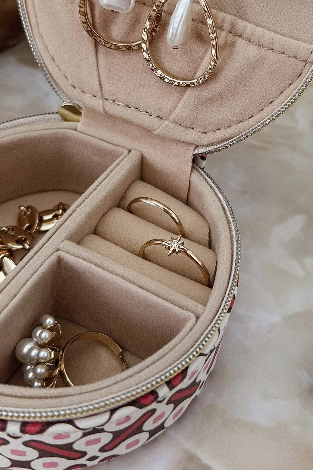 An open round jewelry organizer case with multiple compartments, shown holding several gold rings and pearl-like earrings. The case has a beige interior and a patterned exterior with pink, white, and dark red.