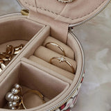 An open round jewelry organizer case with multiple compartments, shown holding several gold rings and pearl-like earrings. The case has a beige interior and a patterned exterior with pink, white, and dark red.