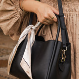 Black handbag with a scarf detail held by a person wearing a beige dress.