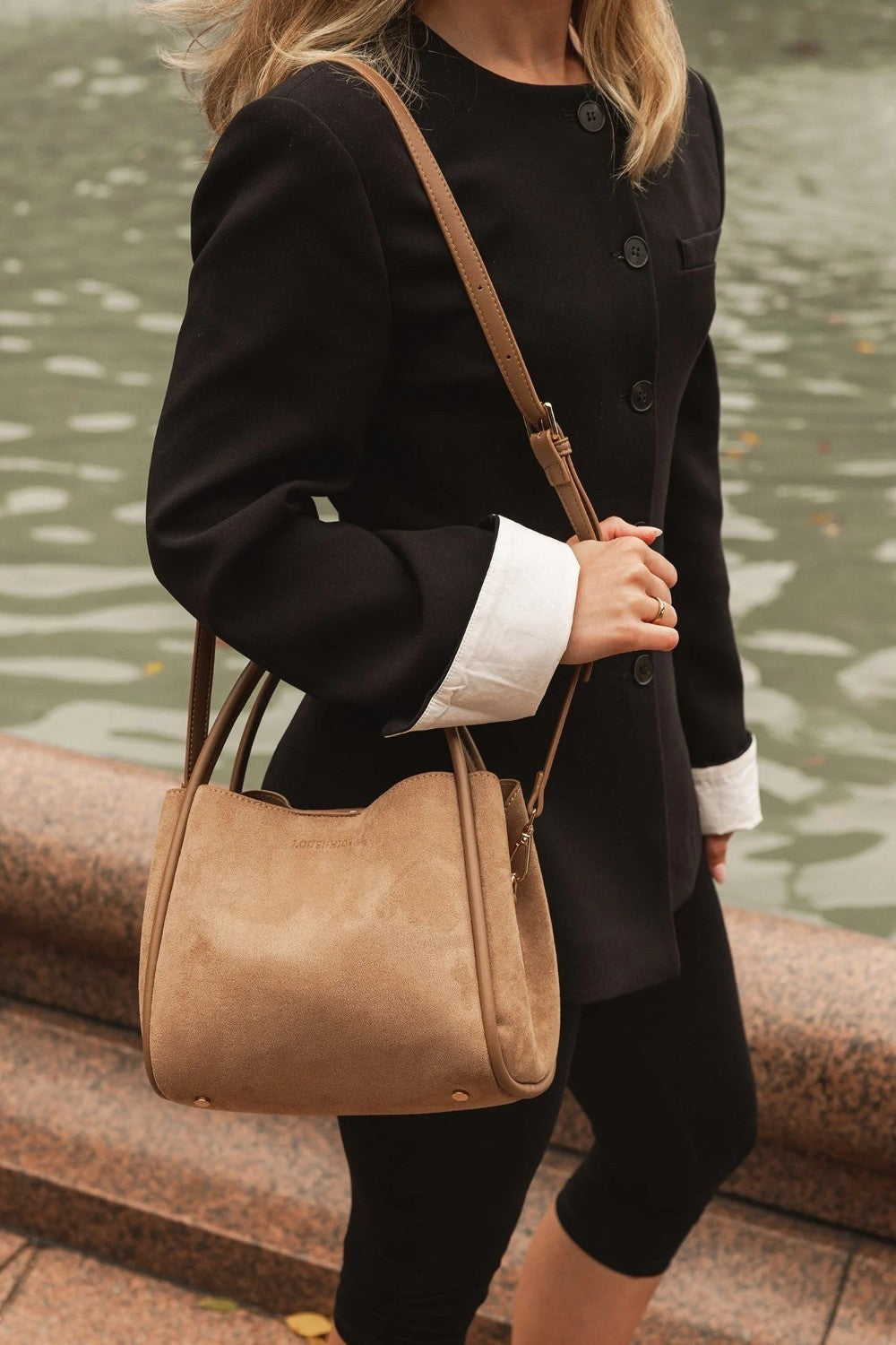 Person wearing a black coat and holding a tan handbag by a body of water.