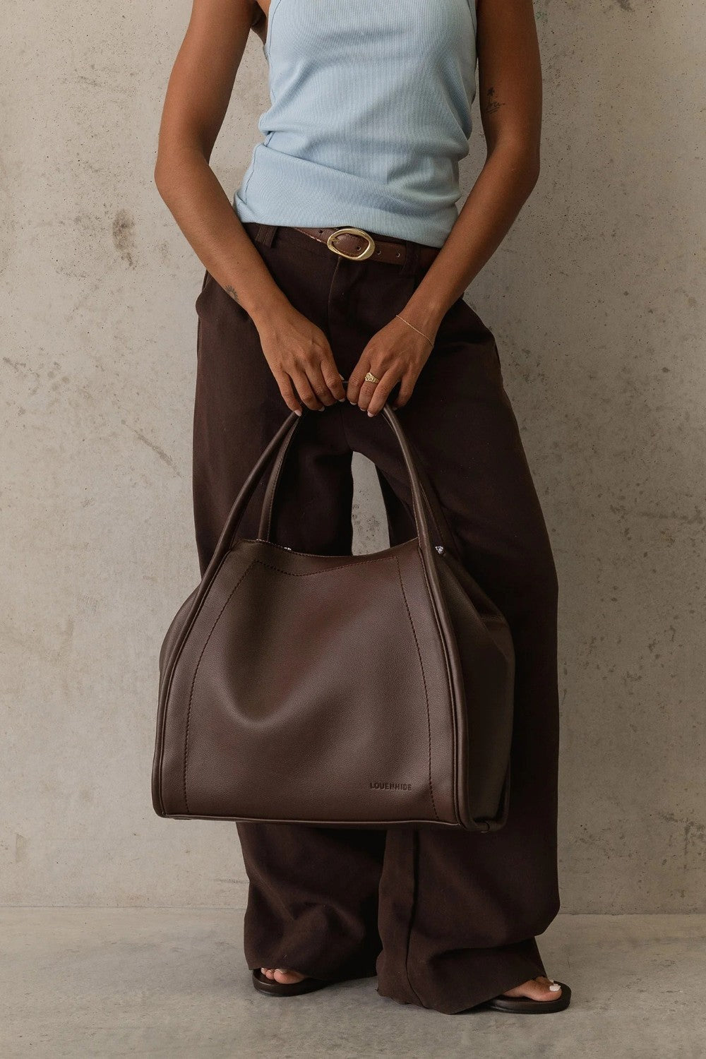 A person wearing dark brown wide-leg pants and a light blue ribbed tank top holds a large dark brown leather hobo handbag against a textured concrete wall.