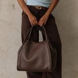 A person wearing dark brown wide-leg pants and a light blue ribbed tank top holds a large dark brown leather hobo handbag against a textured concrete wall.