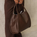A person wearing brown wide-leg trousers and black sandals holds a large brown faux-leather tote bag with double shoulder straps against a textured light gray wall.