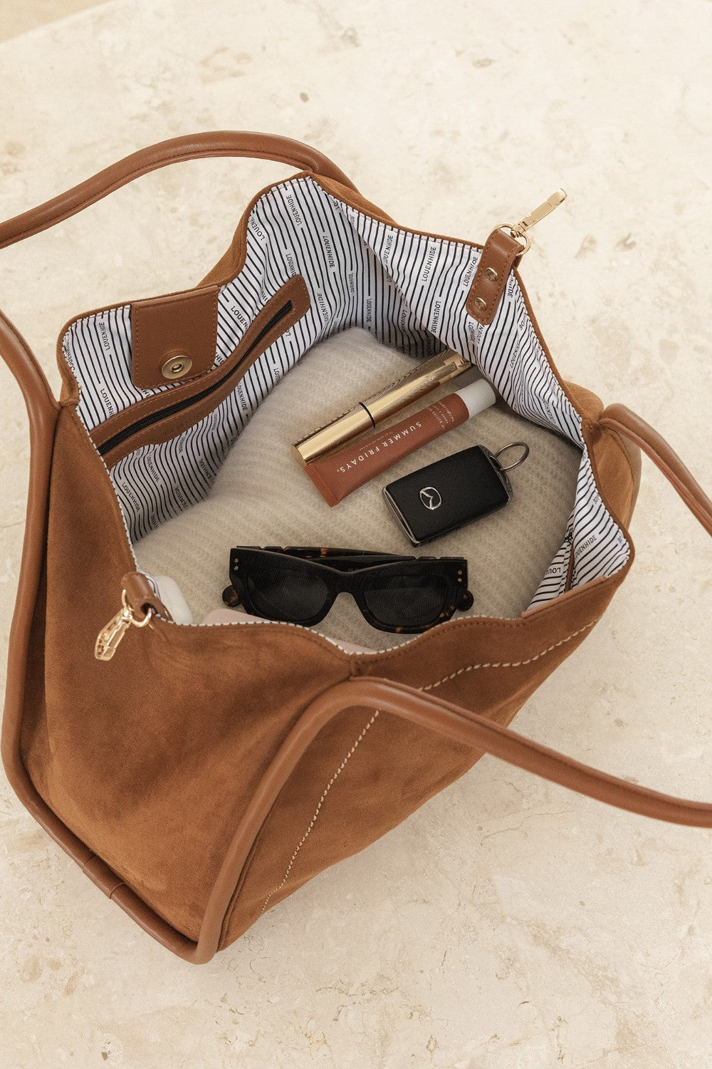 Brown handbag with sunglasses, phone, and lip products on a light background