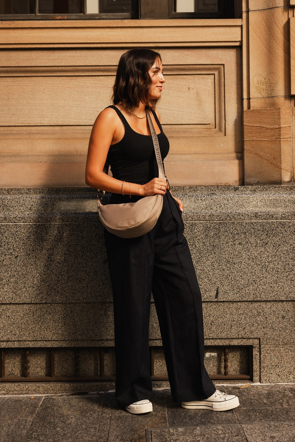 Woman in black outfit with beige bag standing against a building.