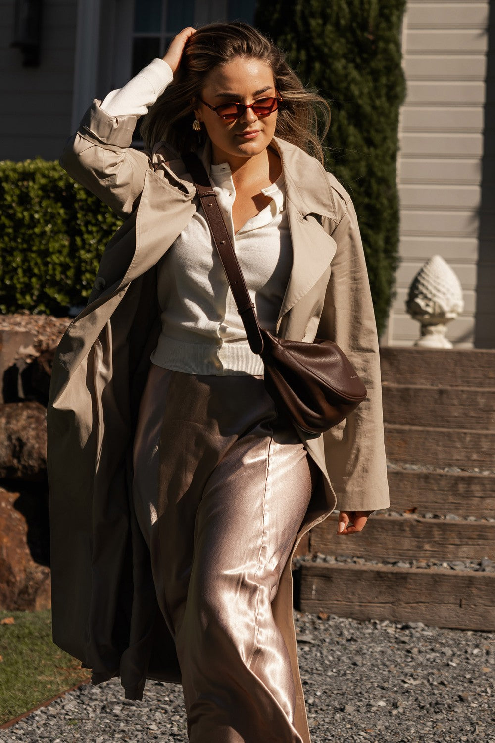 Woman in a beige trench coat and sunglasses holding a brown bag, standing on steps outdoors.
