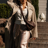 Woman in a beige trench coat and sunglasses holding a brown bag, standing on steps outdoors.