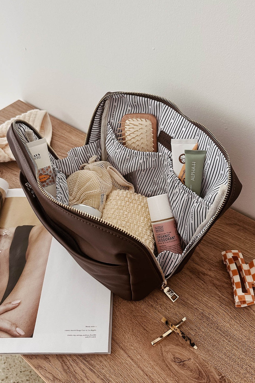 Open brown pleather bag with striped lining containing travel-sized products on a wooden surface.