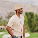 Man wearing a straw hat and beige shirt standing outdoors with mountains in the background
