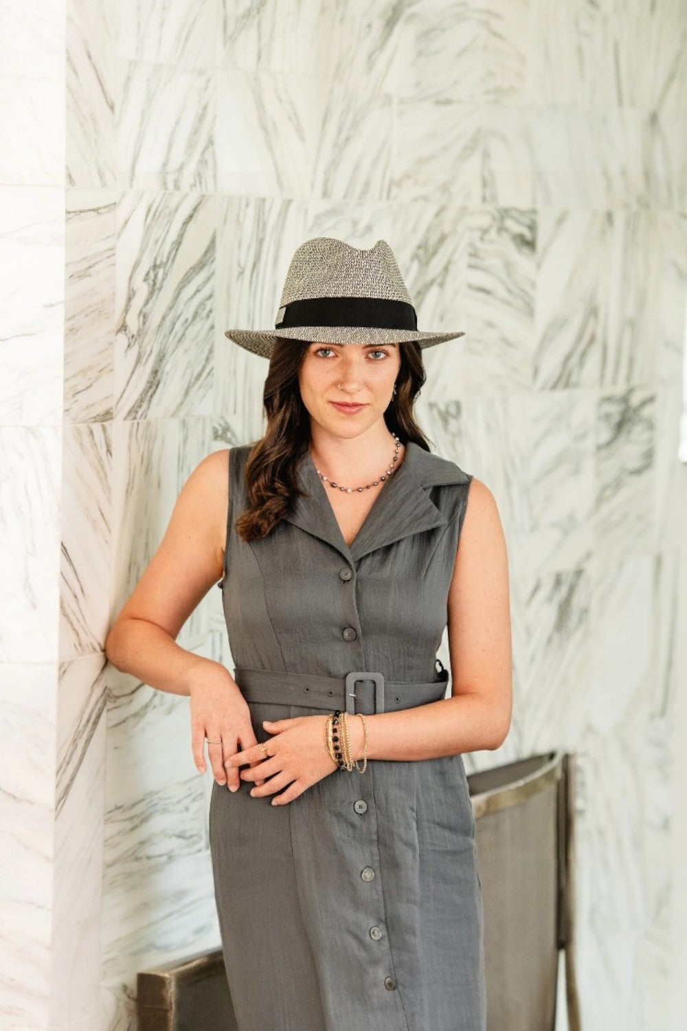 Woman wearing a gray sleeveless dress and hat against a textured white wall.
