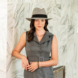 Woman wearing a gray sleeveless dress and hat against a textured white wall.