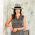 Woman wearing a gray sleeveless dress and hat against a textured white wall.