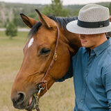 Person in a blue shirt and hat standing next to a brown horse in a grassy field with mountains in the background.