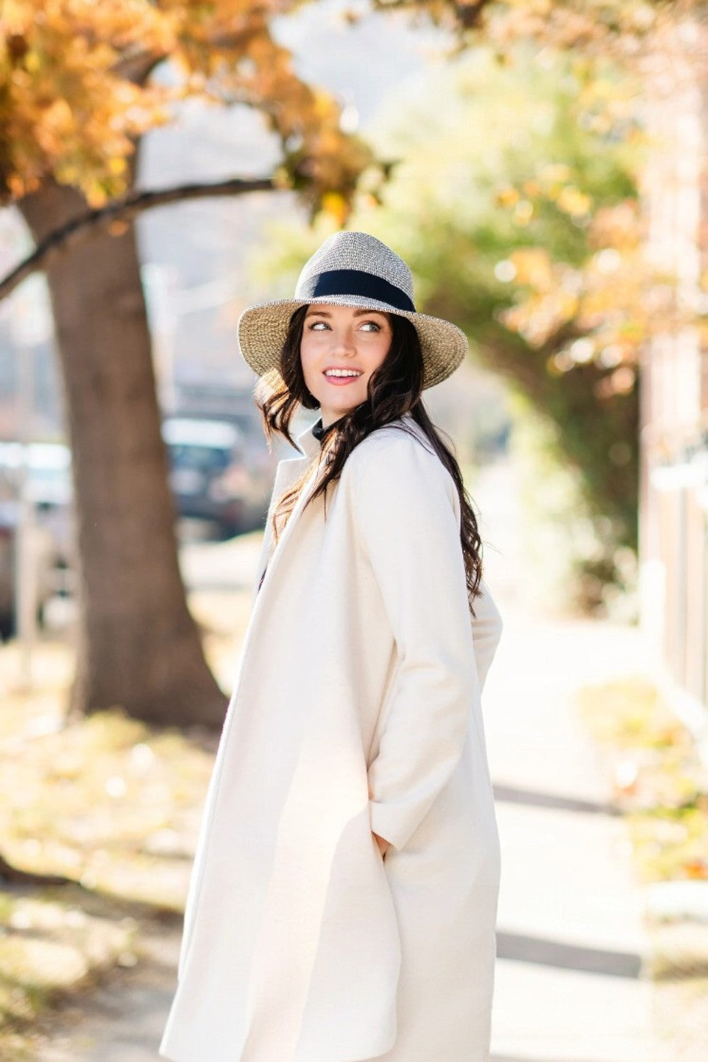 Woman in a long coat and hat standing outdoors with autumn foliage in the background