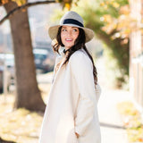 Woman in a long coat and hat standing outdoors with autumn foliage in the background