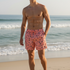 A shirtless man standing on a sandy beach wearing pink swim trunks with a black pattern, with the ocean and sky in the background.