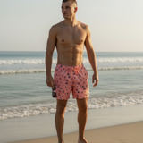 A shirtless man standing on a sandy beach wearing pink swim trunks with a small dark pattern, with ocean waves and a pale sky in the background.