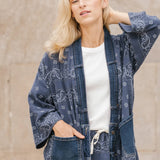 Woman wearing a blue patterned kimono-style garment against a beige wall.
