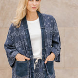 Woman wearing a blue patterned long cardigan over a white shirt against a beige wall.