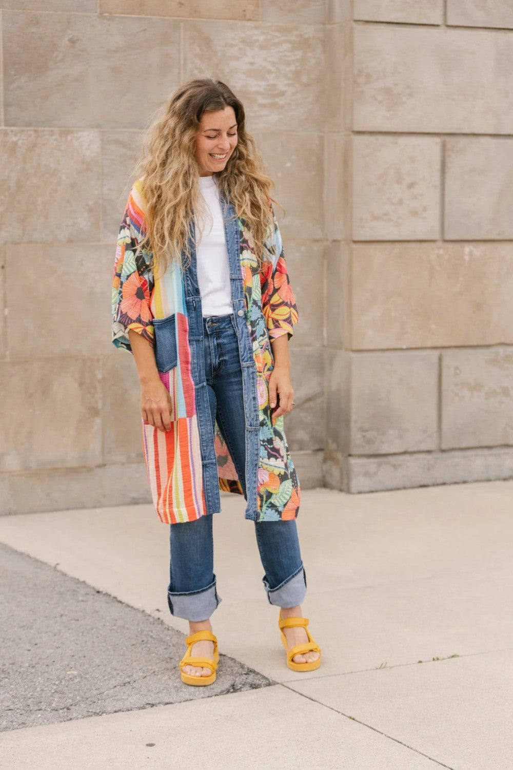 Woman wearing a colorful kimono-style jacket and jeans standing against a stone wall.