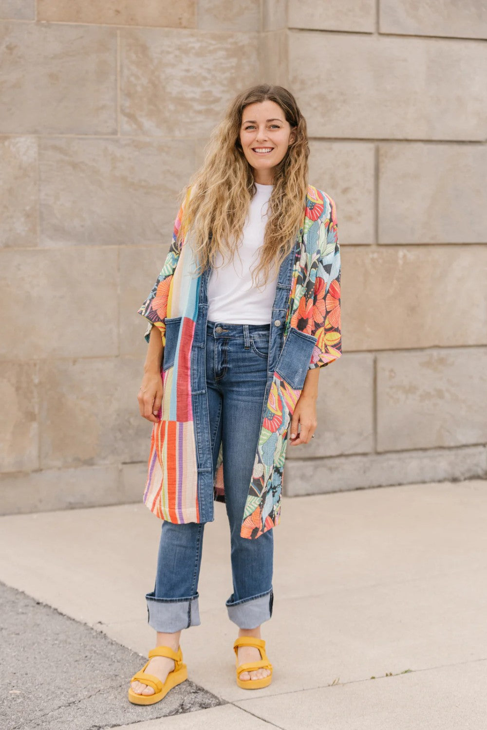 Woman wearing a colorful kimono-style jacket, white shirt, blue jeans, and yellow sandals standing against a stone wall.