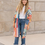 Woman wearing a colorful kimono-style jacket, white shirt, blue jeans, and yellow sandals standing against a stone wall.