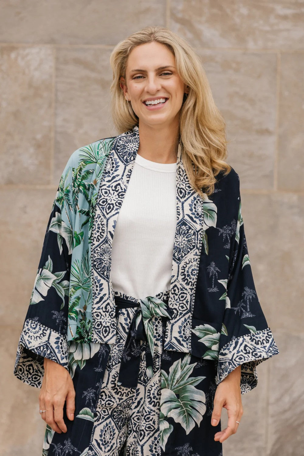 Woman wearing a palm print kimono-style outfit against a stone wall.