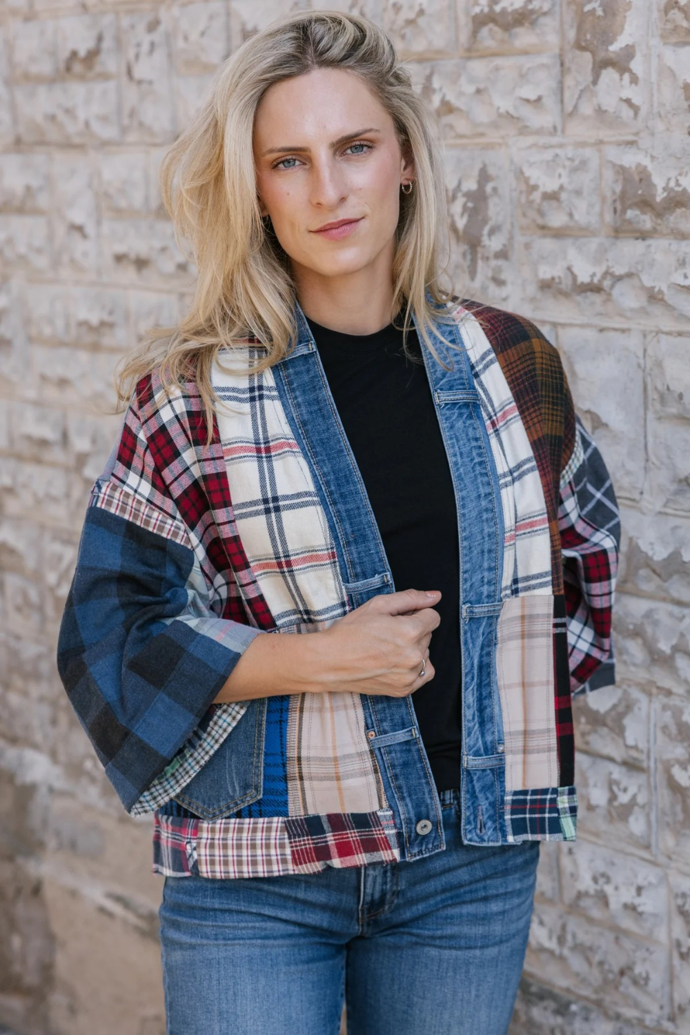 Woman standing in front of a stone wall wearing a black t-shirt and blue jeans with a mixed plaid fabric cardigan with denim trim and pockets.