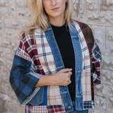 Woman standing in front of a stone wall wearing a black t-shirt and blue jeans with a mixed plaid fabric cardigan with denim trim and pockets.