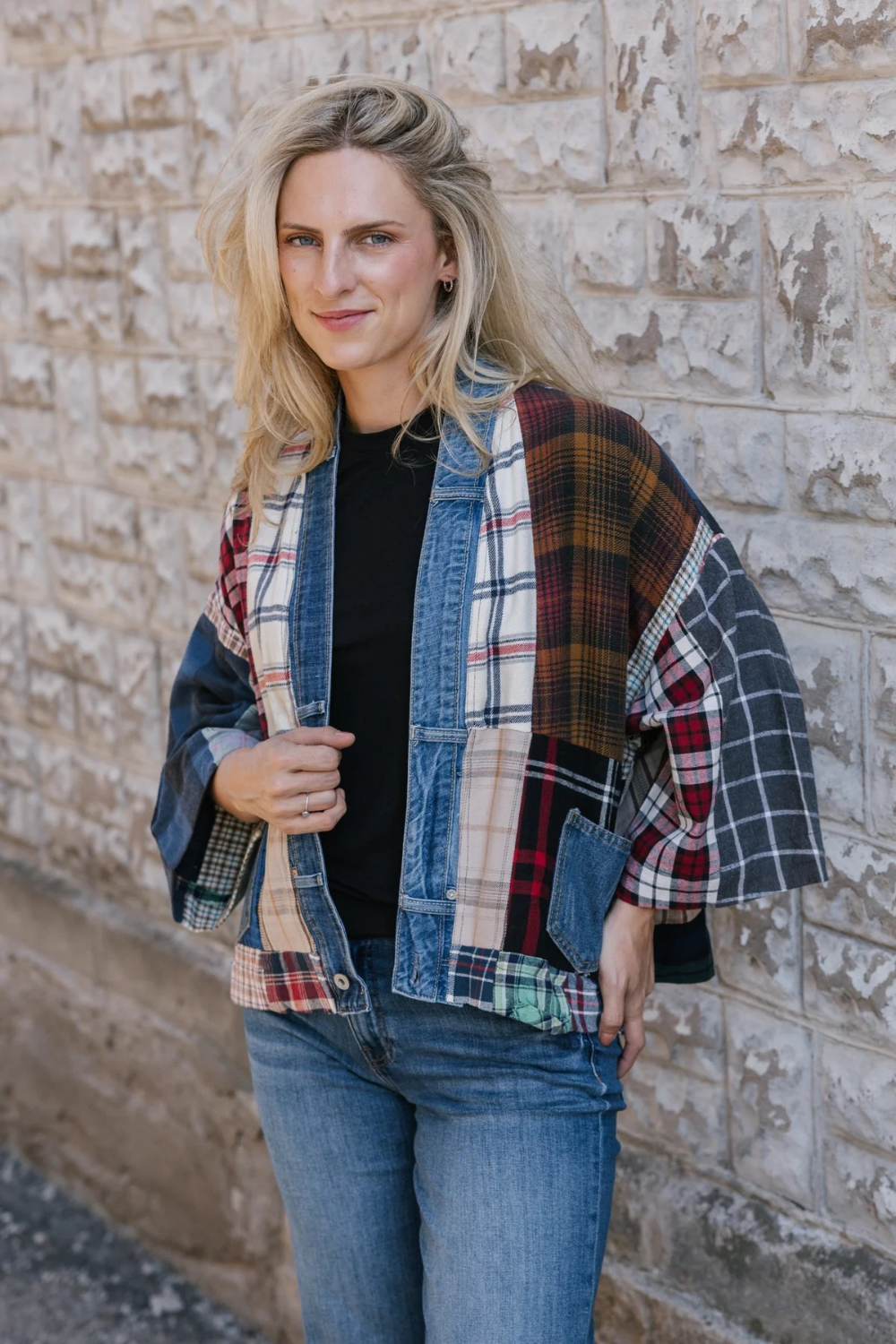 Woman standing in front of a stone wall wearing a black t-shirt and blue jeans with a mixed plaid fabric cardigan with denim trim and pockets.