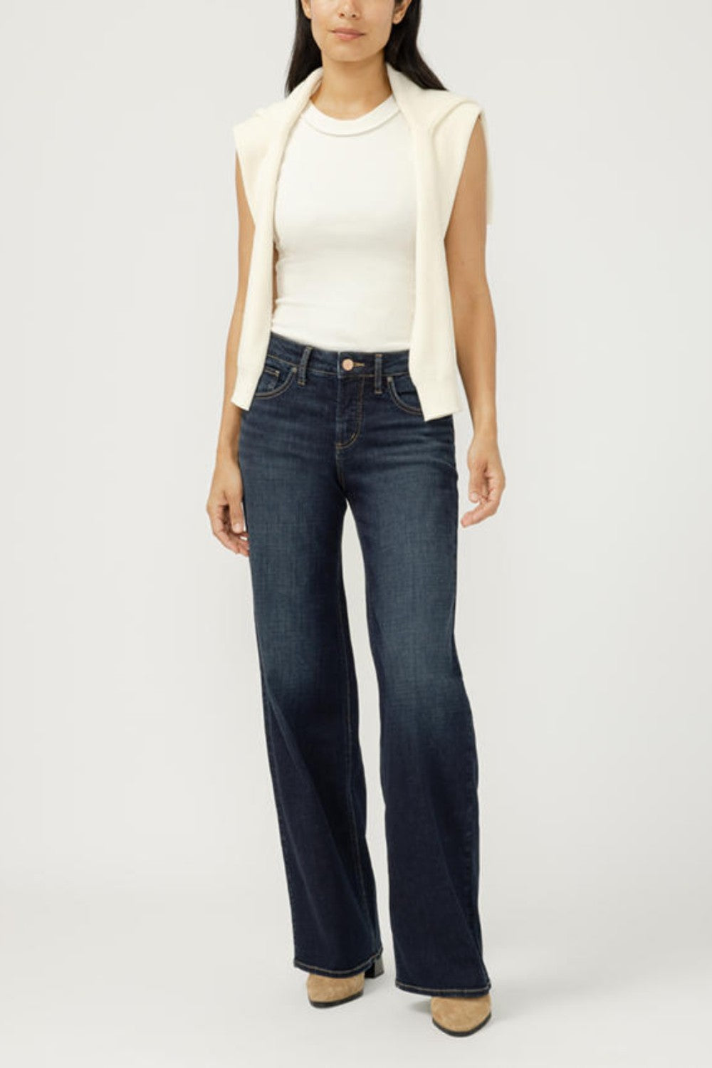 Woman wearing a white top, cream vest, and dark blue jeans on a white background