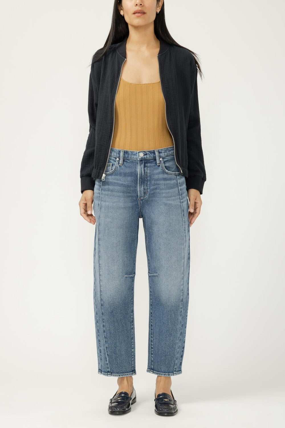 Woman wearing a black jacket, mustard top, and blue jeans on a white background