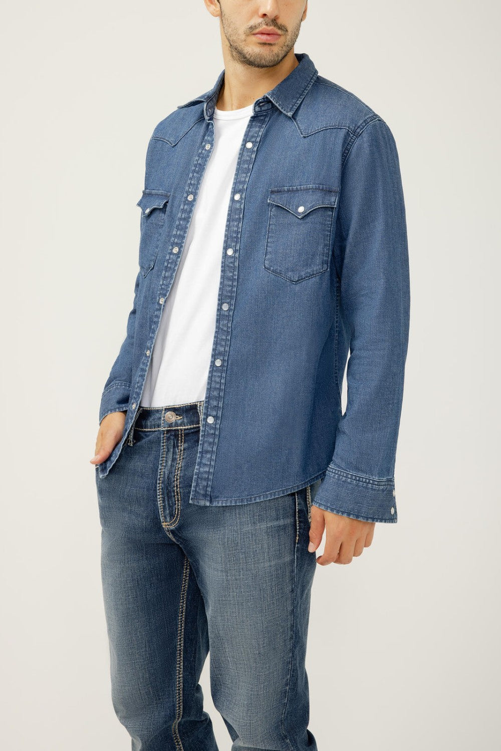 Man wearing a blue denim shirt over a white t-shirt and jeans on a plain background