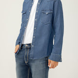 Man wearing a blue denim shirt over a white t-shirt and jeans on a plain background