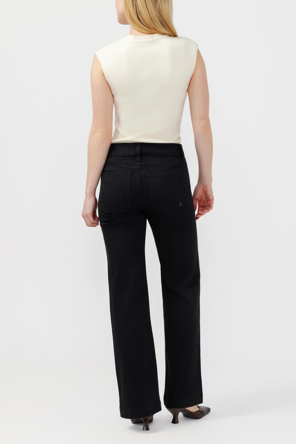 Woman, shown from behind, wearing black jeans with a sleeveless white top and dark shoes against a light backdrop.