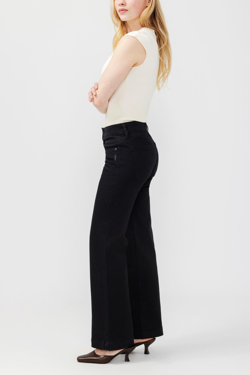 Woman wearing black jeans with a sleeveless white top and dark shoes against a light backdrop.