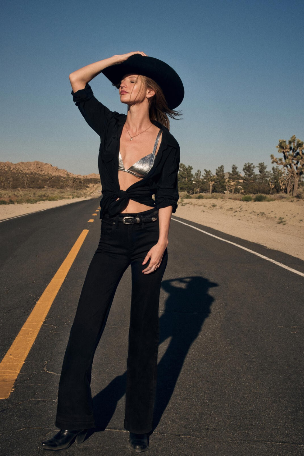 Woman standing in an outdoor setting with a black cowboy hat, a black cardigan sweater with a silver coloured bra revealed, black jeans with a black belt and black boots.
