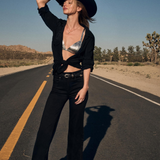 Woman standing in an outdoor setting with a black cowboy hat, a black cardigan sweater with a silver coloured bra revealed, black jeans with a black belt and black boots.
