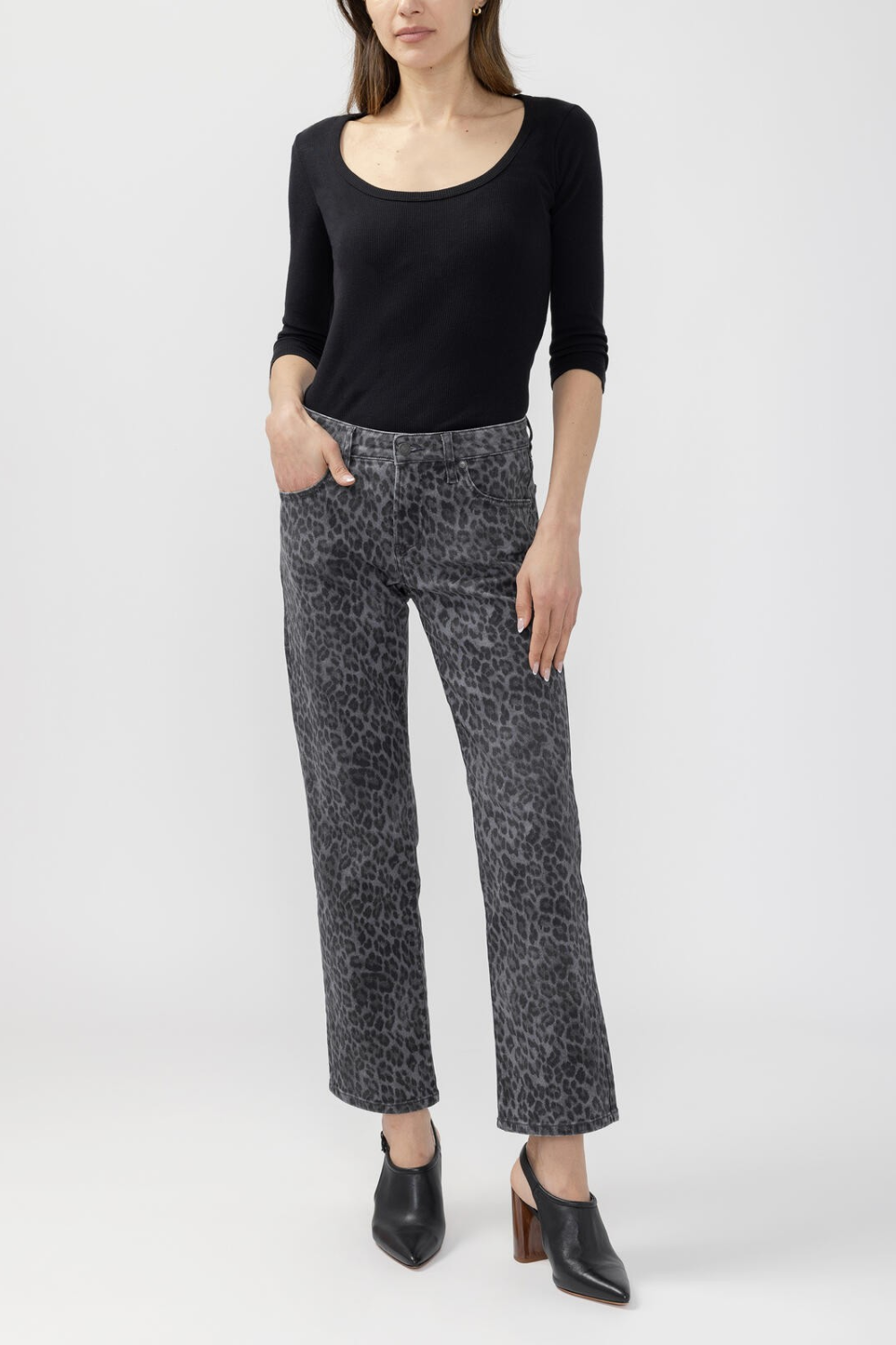 A woman modeling a pair of cropped black and gray leopard-print pants, paired with a black long-sleeve scoop-neck top and black mule heels, against a light gray studio background.