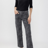 A woman modeling a pair of cropped black and gray leopard-print pants, paired with a black long-sleeve scoop-neck top and black mule heels, against a light gray studio background.