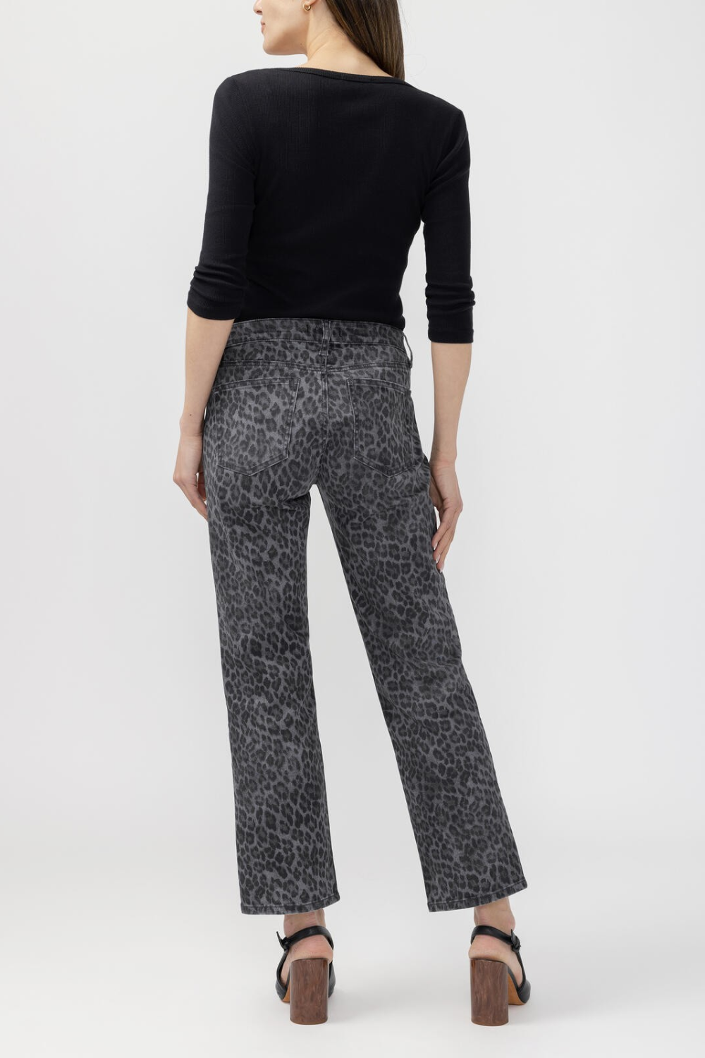 A woman, shown from behind, modeling a pair of cropped black and gray leopard-print pants, paired with a black long-sleeve top and black mule heels, against a light gray studio background.