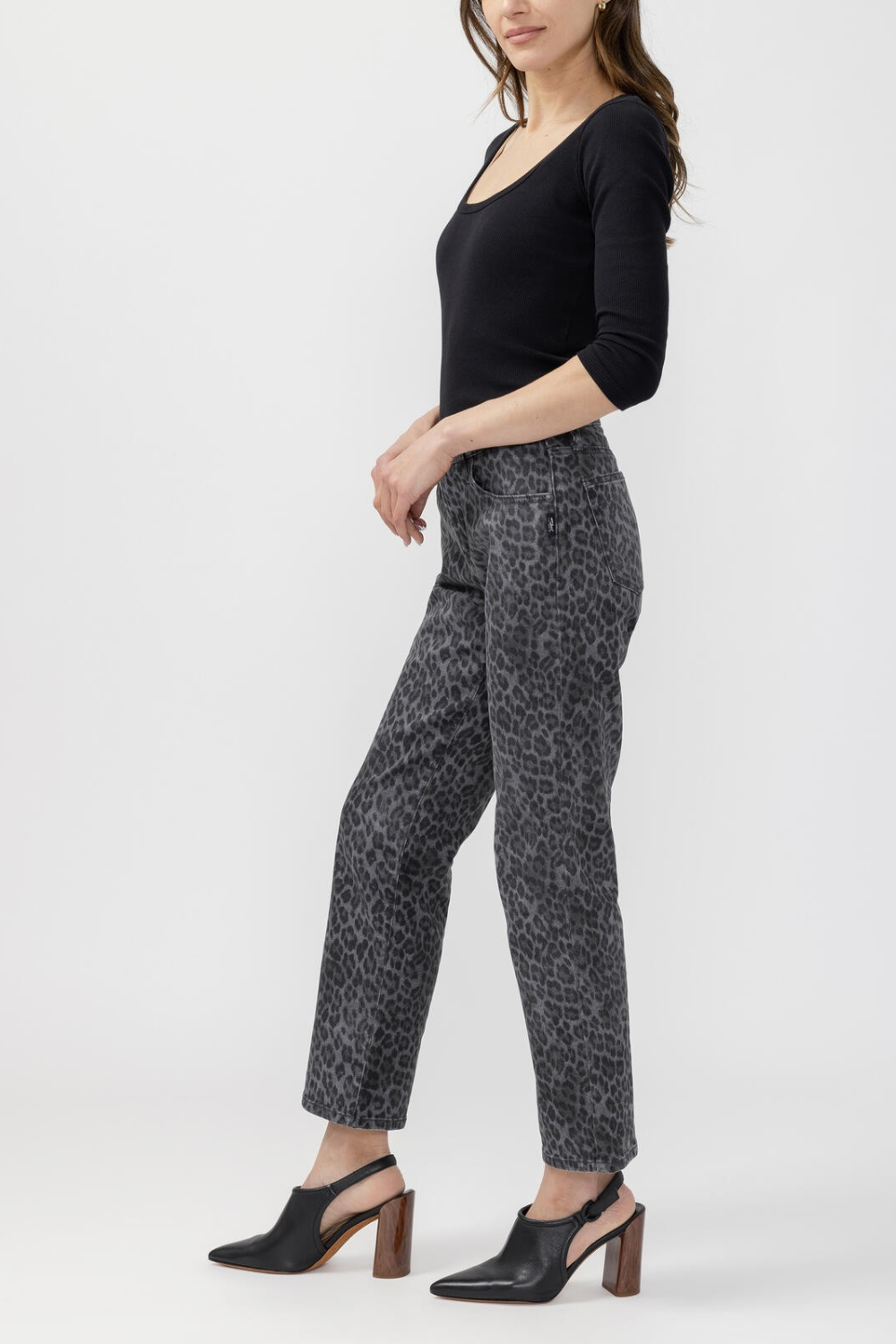 A woman modeling a pair of cropped black and gray leopard-print pants, paired with a black long-sleeve scoop-neck top and black mule heels, against a light gray studio background.