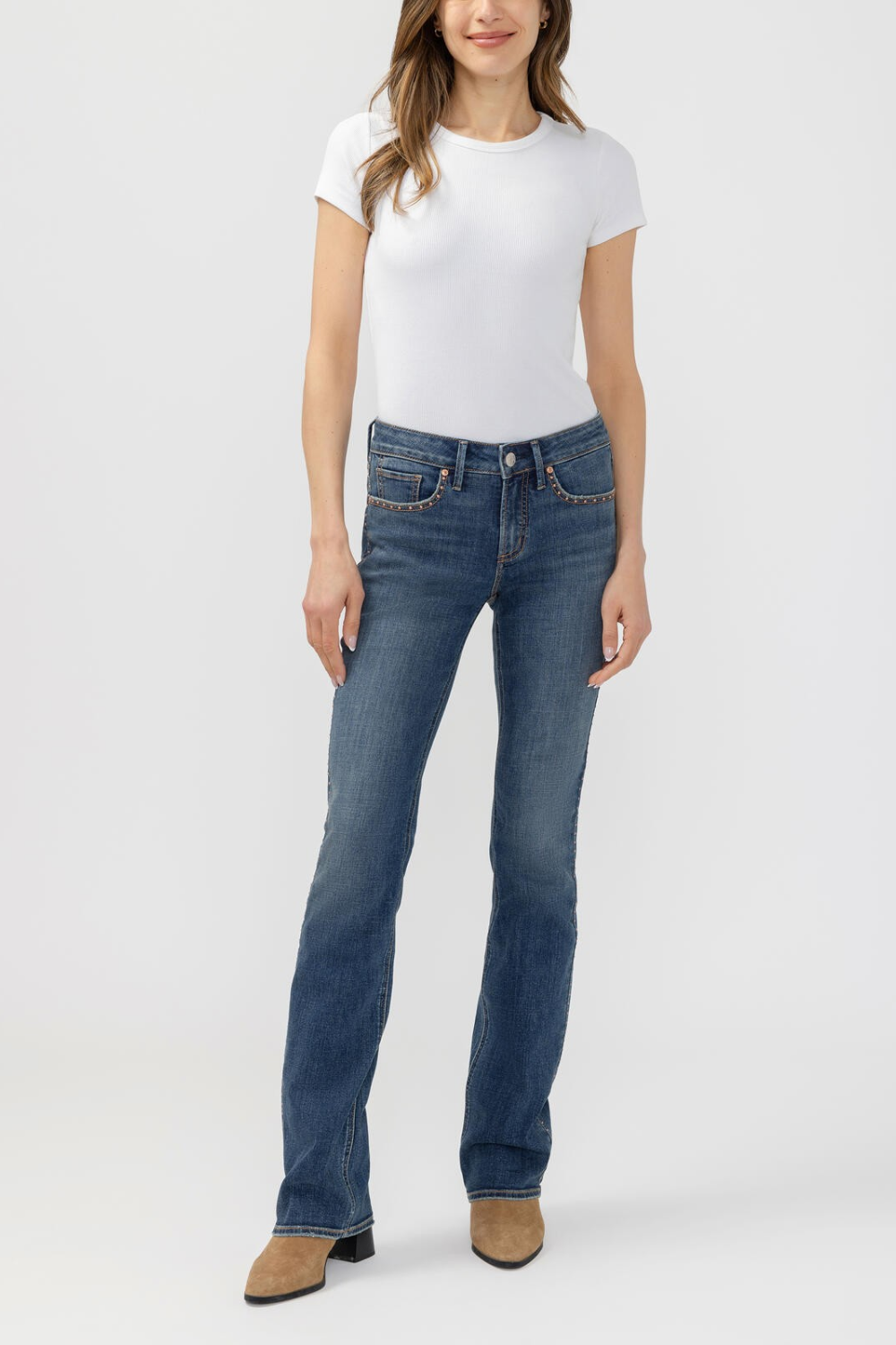 A woman modeling a pair of medium-wash blue jeans with a white t-shirt and brown ankle boots, standing against a light gray studio background.