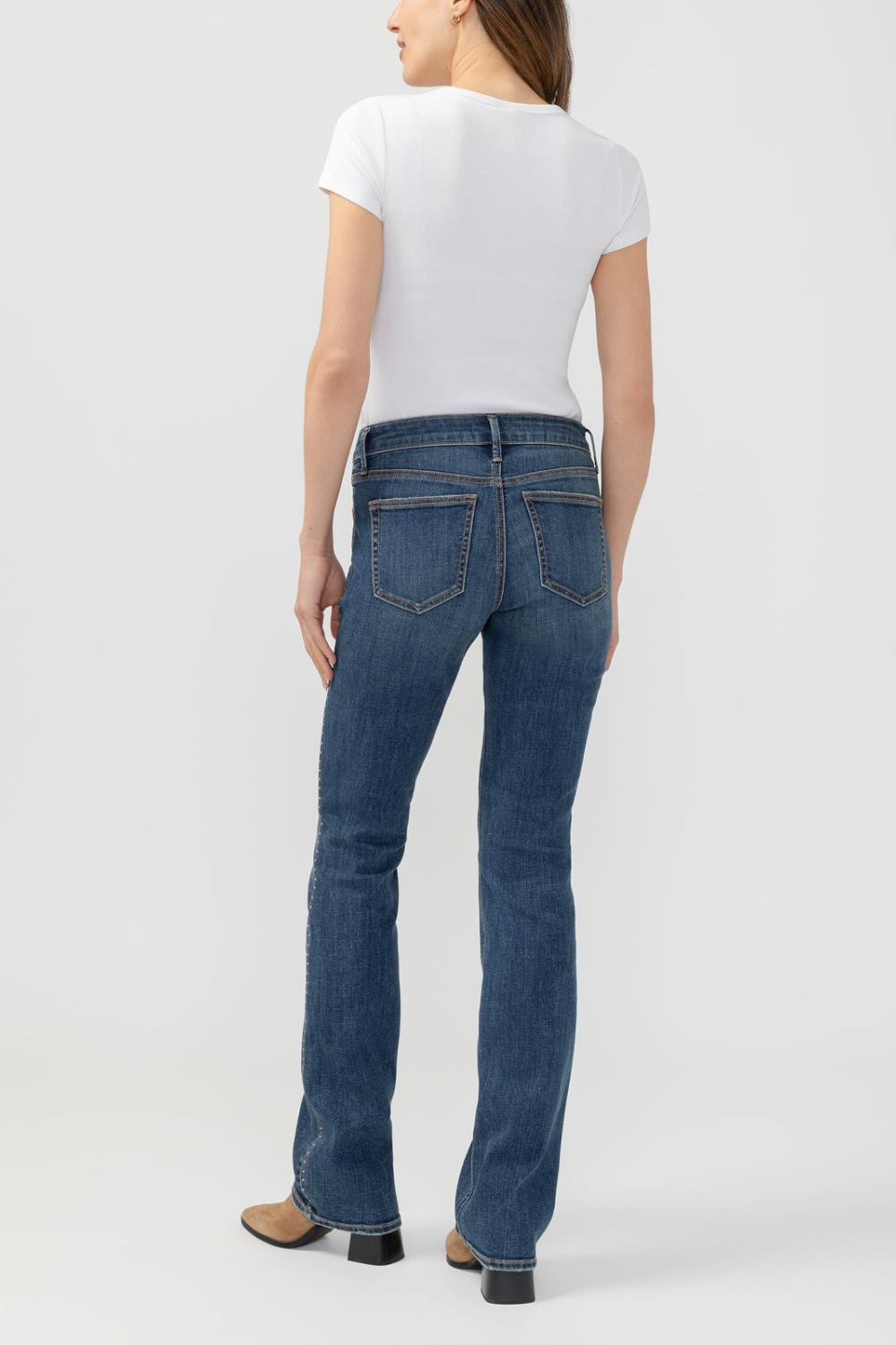 A woman shown from the back wearing blue flare jeans, a white short-sleeve top, and brown heeled shoes against a light gray studio background.
