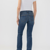 A woman shown from the back wearing blue flare jeans, a white short-sleeve top, and brown heeled shoes against a light gray studio background.